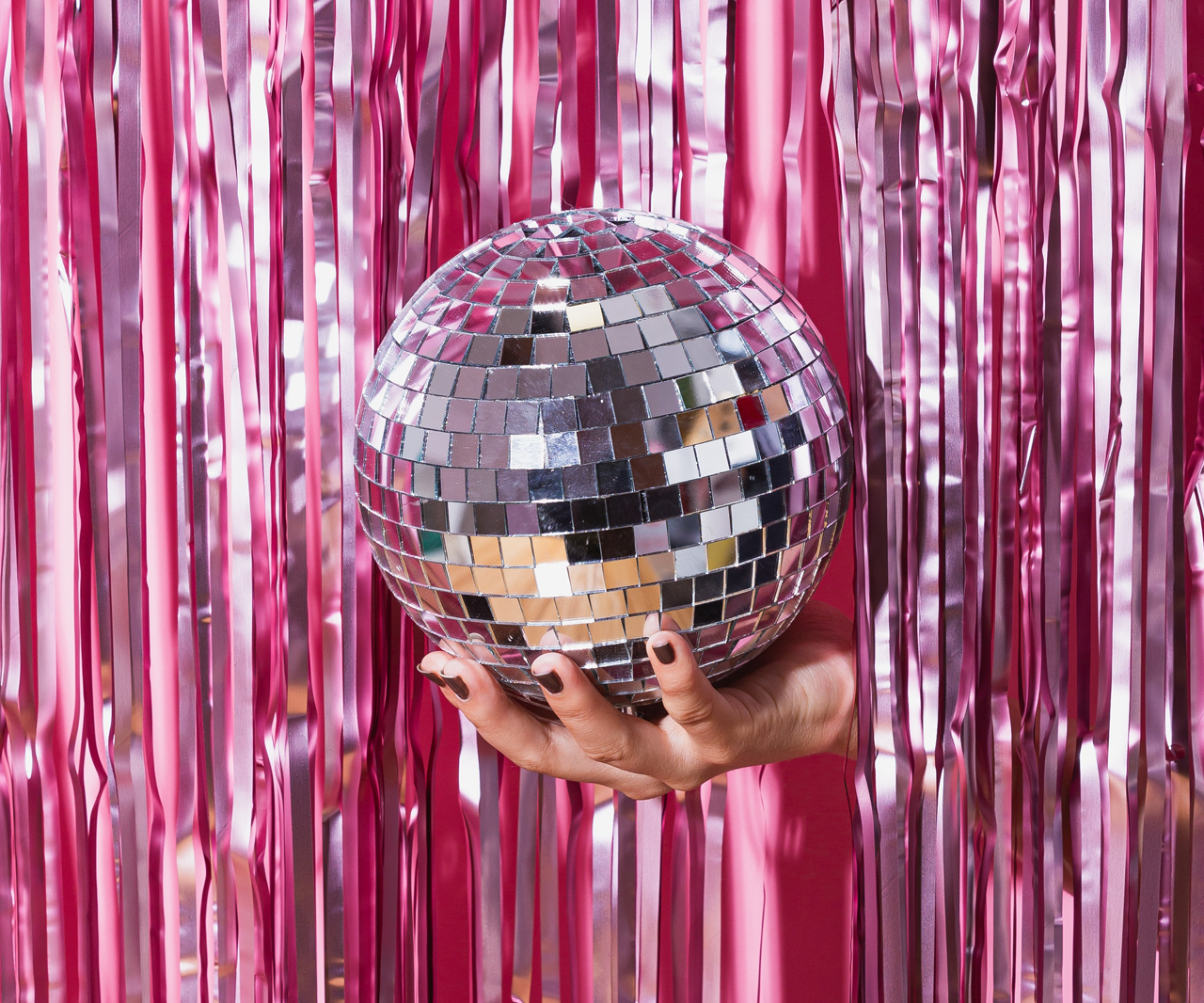 Hand holding a disco ball in front of a pink tinsel backdrop.