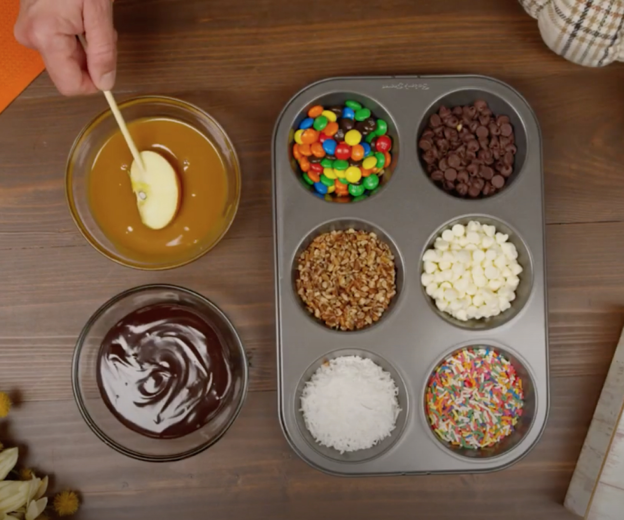 Cupcake tin filled with different toppings and someone dipping an apple into caramel.