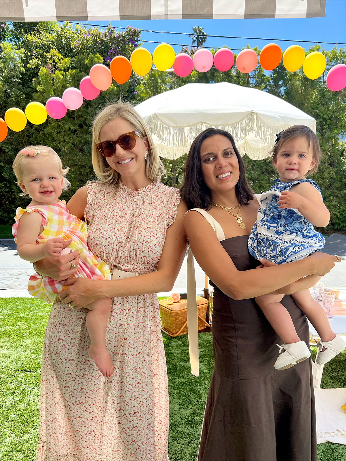 Two moms holding their daughters under a colorful balloon arch.