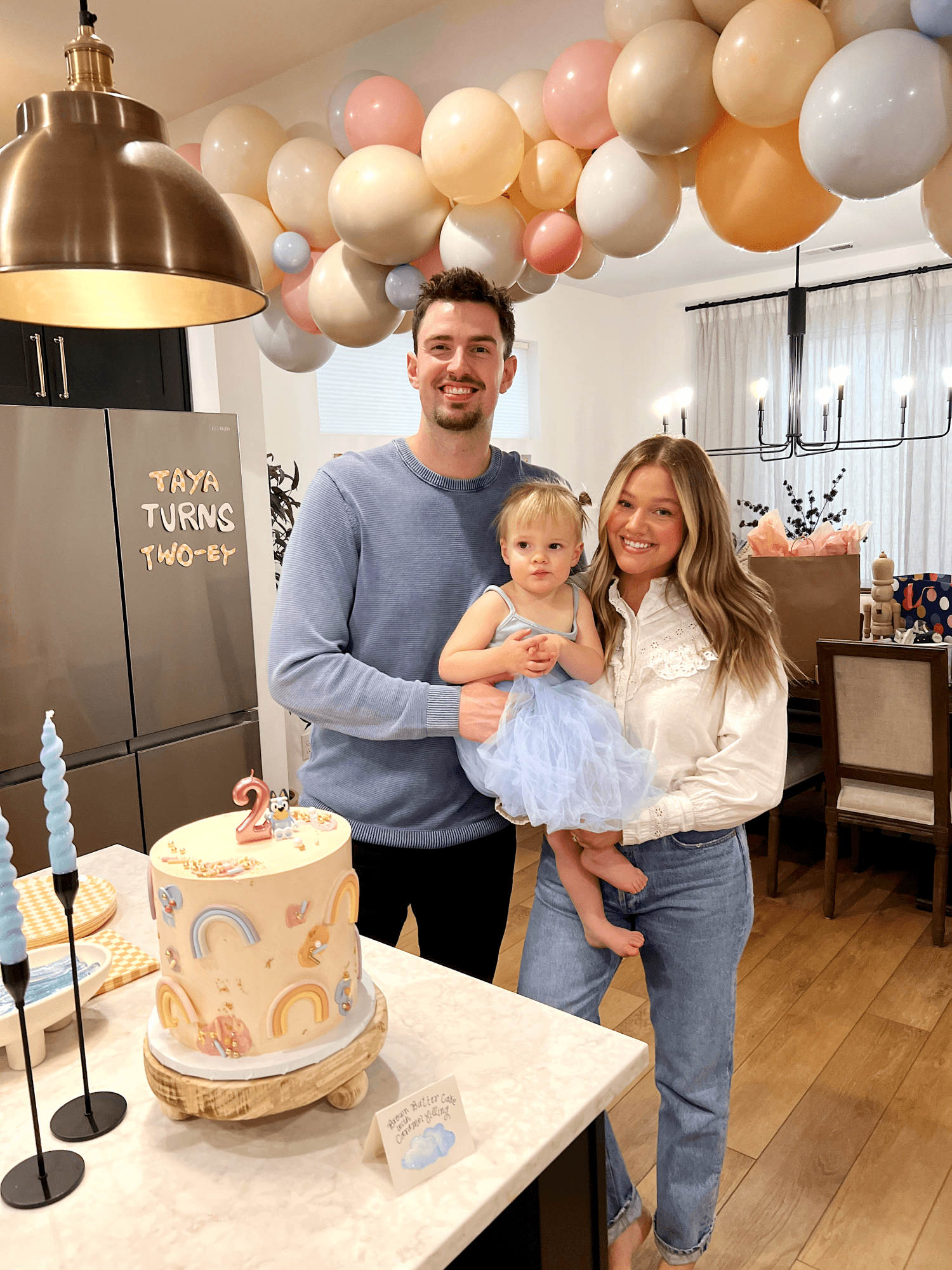 Taya and her parents smiling next to the cake with balloons in the background.