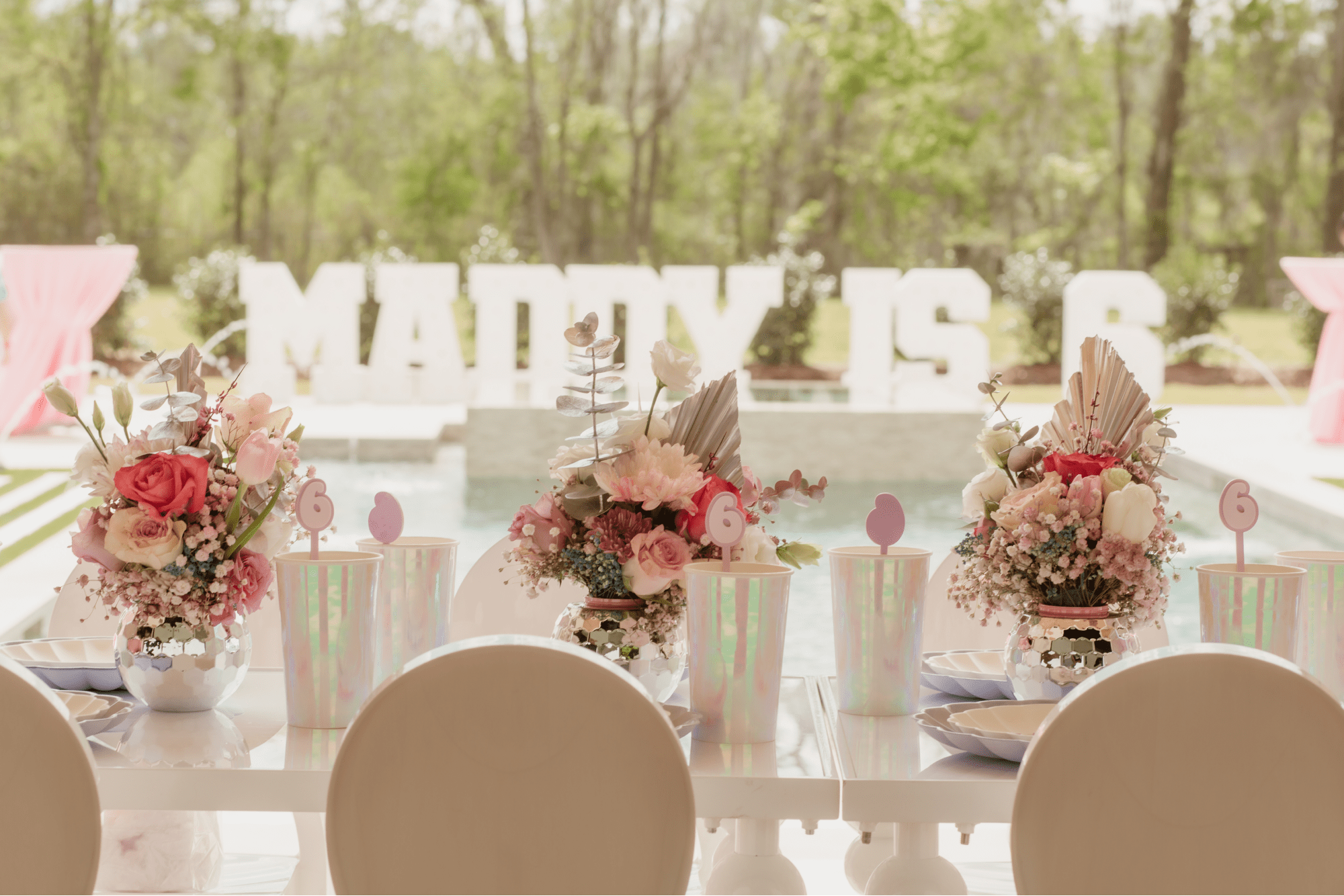 Table set with metallic cups and vases of flowers. A sign saying "Maddy is 6" can be seen in the background.