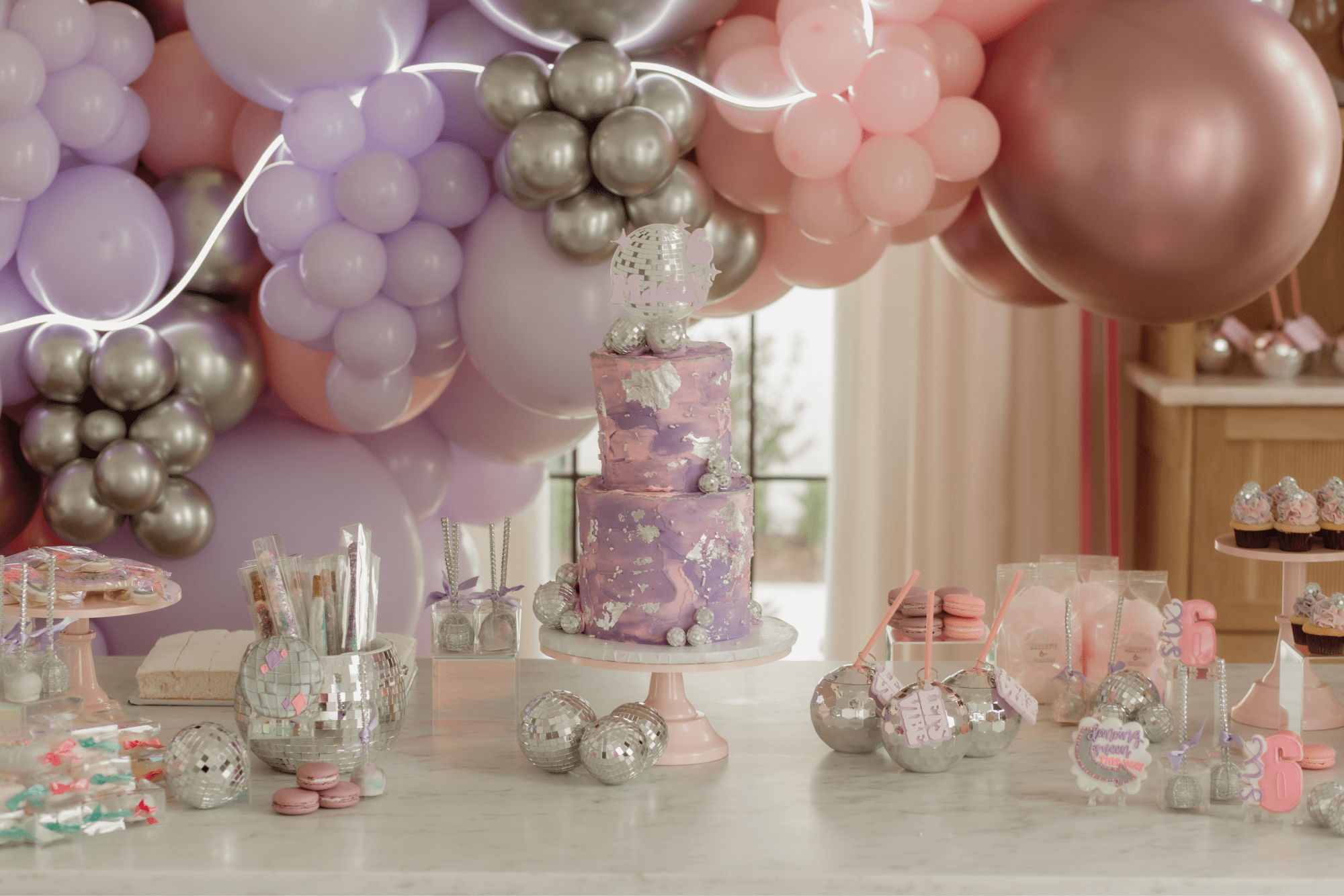 The birthday cake on a table with disco ball cups and miniature cupcakes.