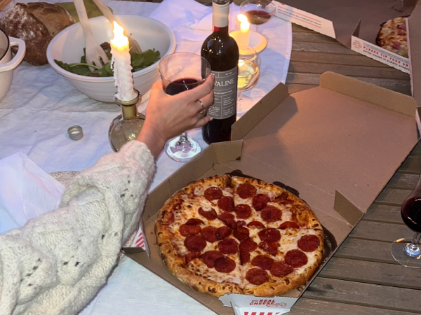 An outreached hand holding a glass of red wine next to a pepperoni pizza.