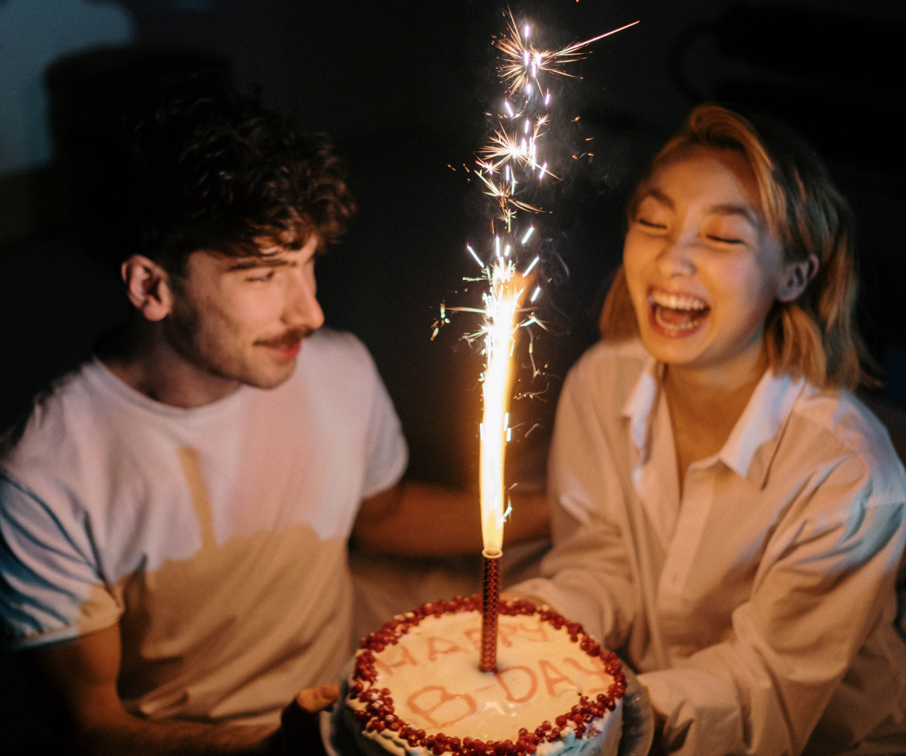 A man passing a woman a birthday cake with a sparkler.