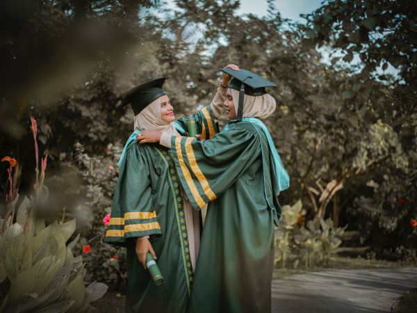 Two graduates helping each other with their sashes and caps.