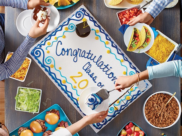 Overhead shot of people cutting a graduation party cake and eating tacos and burgers.